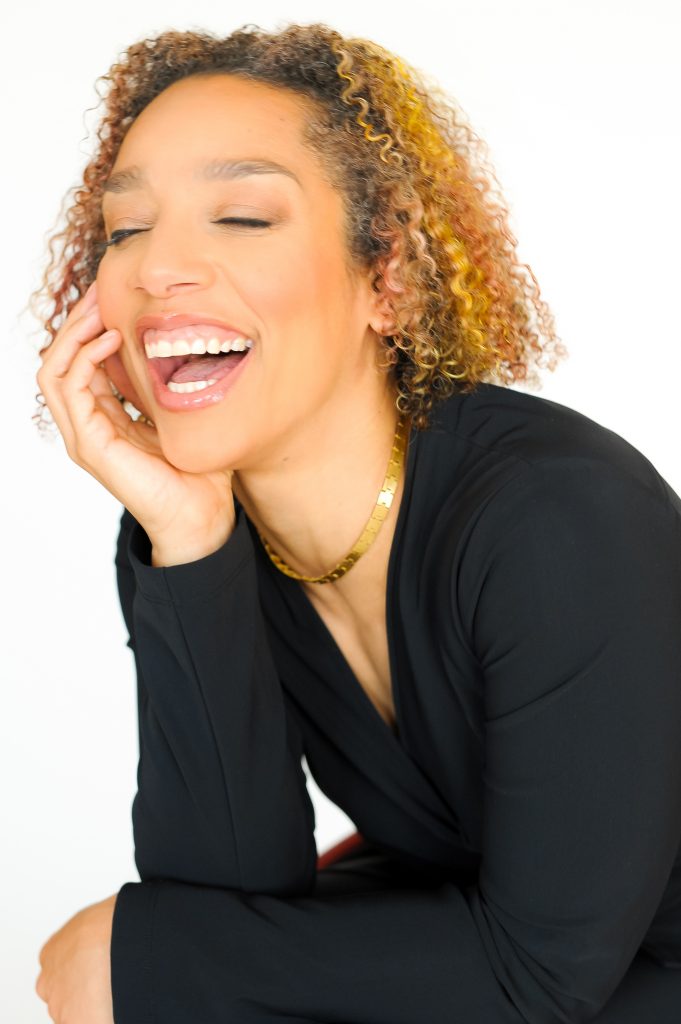 A Black woman leans forward and smiles brightly, teeth showing and eyes closed, as she rests her chin in her palm. She has light brown skin, curly shoulder-length hair with subtle highlights, and wears a black blouse and sleek gold necklace.