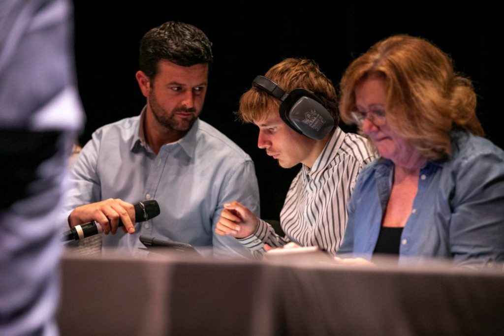 Photo from event, Included in the Picture, a panelist wearing noise-cancelling headphones looks down at their tablet. The panelist to their right is holding up a mic in front of them.