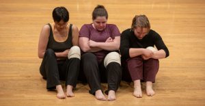 Photo from the Dancing Disability 2019 Open Studio. Three dancers sit next to each others, their knees bent and arms wrapped across their body. They each wear a white piece of fabric, wrapped like a bandage, on their left knee.