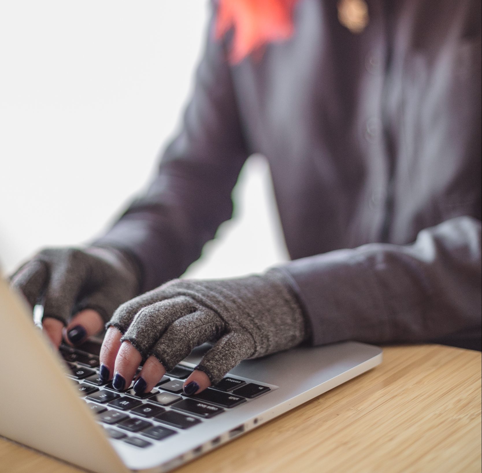 An Asian disabled woman types on a laptop while wearing compression gloves. The hands and keyboard are the focal point.