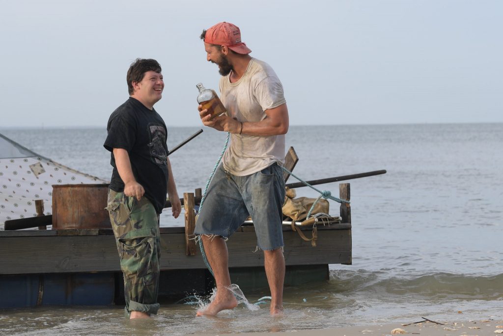 Still from the film Peanut Butter Falcon of stars Zack Gottsagen, an actor who has Down's syndrome, and Shia LaBeouf. They are standing in the ocean, ankle-deep, facing each other, and laughing.
