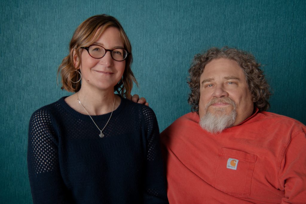 Crip Camp producers Nicole Newnham and James LeBrecht pose against turquoise background, they are both grinning.
