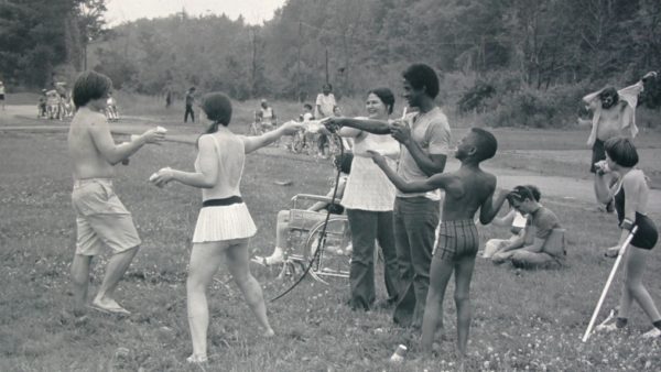 Black and white image of youth at Camp Jened, they are gathered on a grassy field. Three campers reach towards a hose that is spraying water. Several campers have on bathing suits.