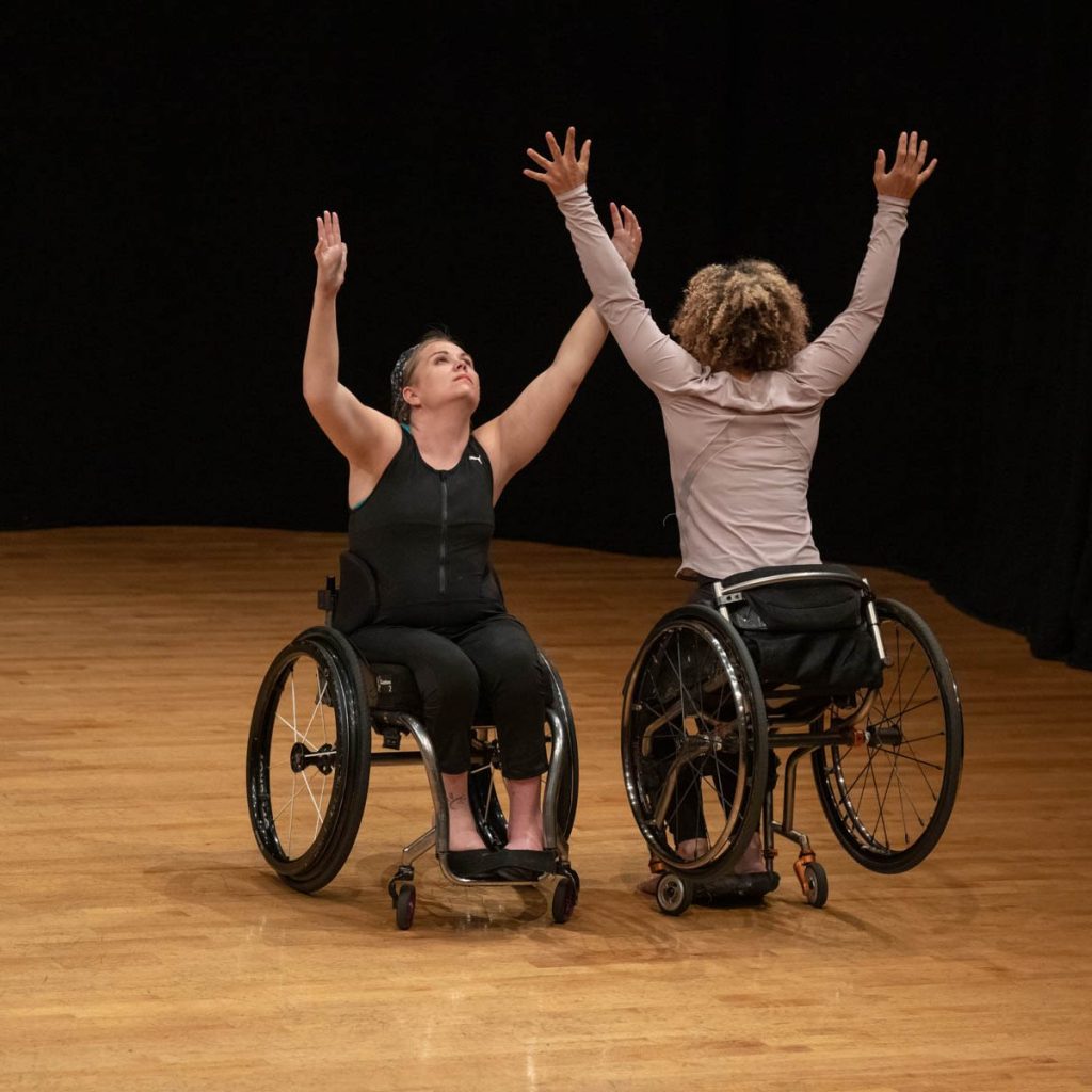 Photo of two dancers, Harmanie Taylor and Alice Sheppard, both using wheelchairs. Their face each other, their arms reaching up. Harmanie looks up to the ceiling, and Alice, whose back is to the camera, leans forward, lifting her wheelchair's wheels a few inches off the ground.