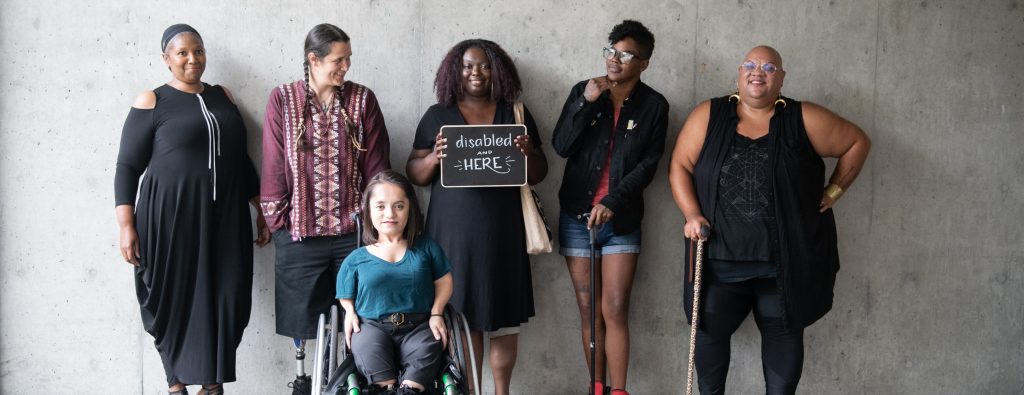 Six disabled people of color smile and pose in front of a concrete wall. Five people stand in the back, with the Black woman in the center holding up a chalkboard sign reading "disabled and here." A South Asian person in a wheelchair sits in front.