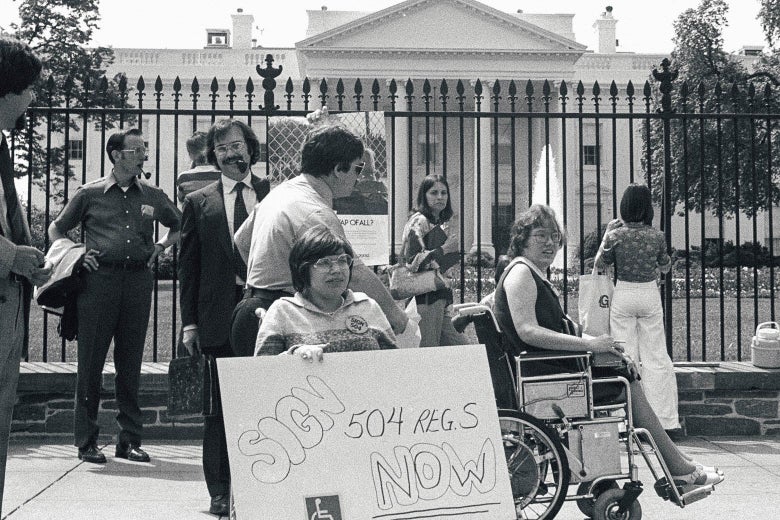Black and white image of protesters, the two in the front are using wheelchairs and one holds a sign that reads Sign 504 Regs Now