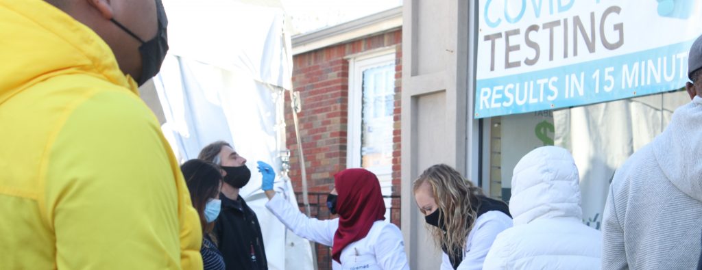 An outdoor COVID-19 testing site. A practitioner wearing a hijab and white lab coat swabs the nose of a community member.