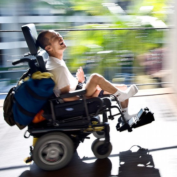 Tim Jin smiling and moving down an outdoor hallway in their power-wheelchair.