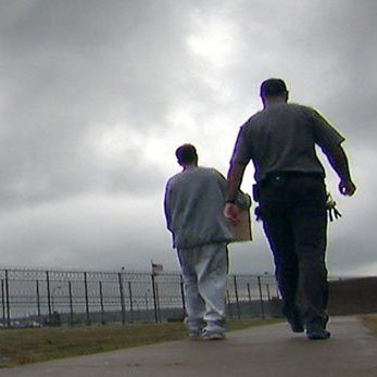Two people walk away, their backs to the viewer. One individual is wearing grey sweatpants and sweatshirt and carrying a box. The person beside them is wearing a beige uniform with a large ring of keys attached to their belt.