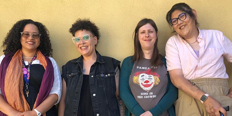 4 disabled femmes stand before a yellow stucco wall. From Left to Right: Sade is a light skinned Black femme with shoulder length ringlets wearing orange and plum scarves over a black dress and white blouse. Tasha is a light skinned Black femme with a short â€˜do with cropped sides wearing a denim vest over a long black dress. System Hatfield is the Jewish femme with shoulder length brown hair wearing a cardigan, clown tee, and skirt. Lilac is the Latinx femme with a pink shirt, shorts and blue cane. All smile gleefully.