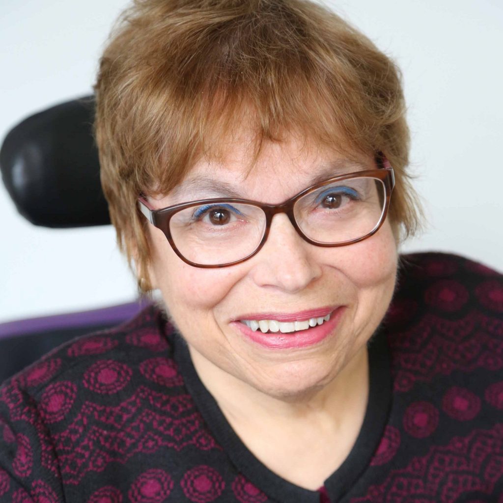 A headshot of Judy Heumann, a cis-gender white woman who is a wheelchair user with short brown hair. She is wearing brown glasses, a maroon and black embroidered cardigan with the top buttoned, and a matching maroon shirt. She is smiling kindly.