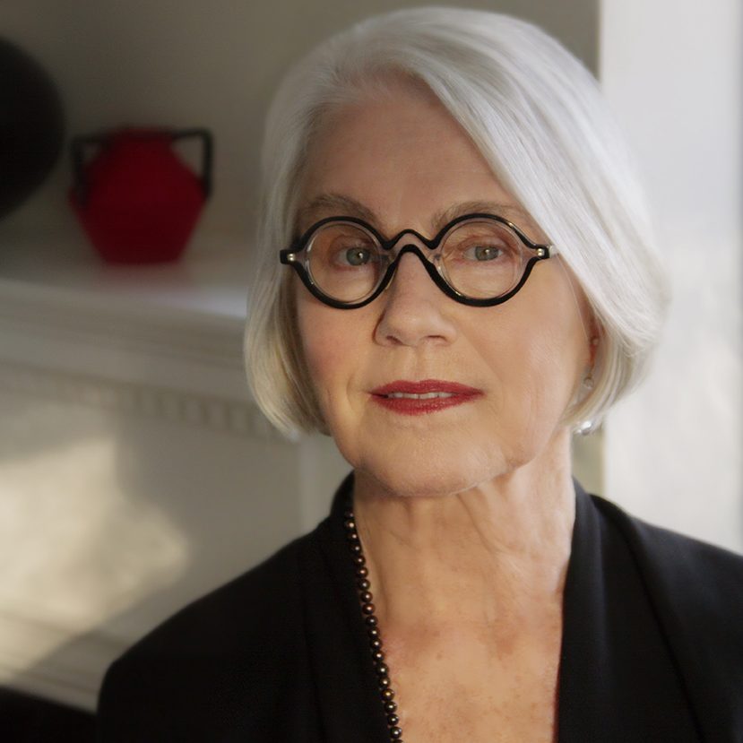 Portrait of Rosemarie Garland-Thomson. She has silver hair styled in a neat, short bob, and smiles gently to the camera. She wears eccentric, black-rimmed, round glasses.