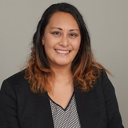 Portrait of Juliann Anesi smiling against a light grey background. She is wearing a black blazer and black and white patterned shirt.