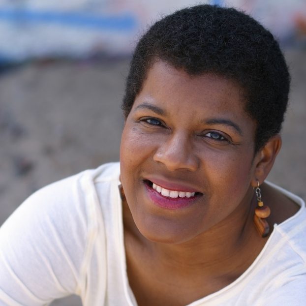 Tananarive Due, a Black woman, smiles upward toward the camera wearing fuchsia lipstick, a white long-sleeved shirt, and dangly wooden earrings.