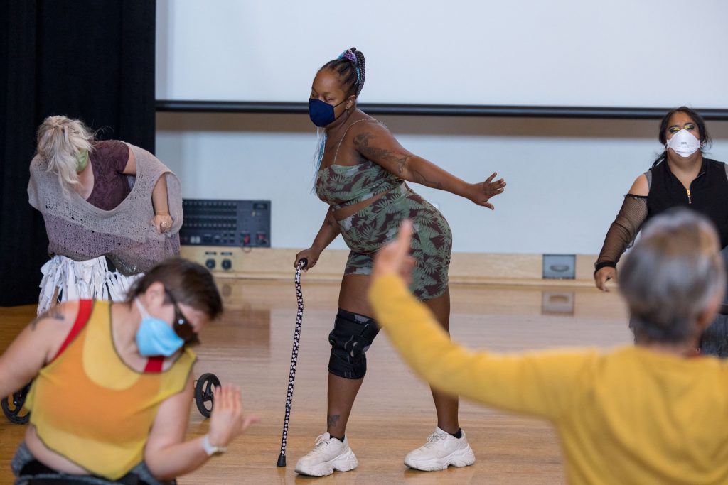 Photo of five members of the Dancing Disability Lab captured in motion while moving. The center of the photo shows Saira Barbaric standing with their cane in a leaning position and left hand arm extended out.