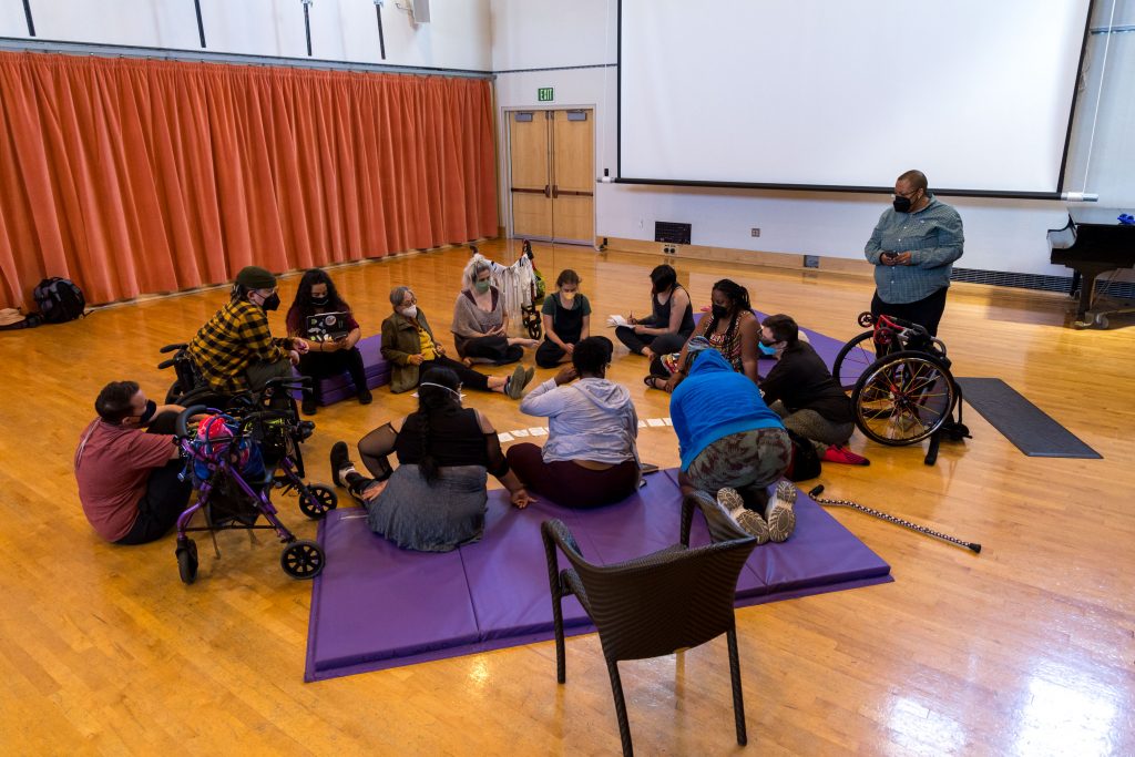 2022 Dancing Disability Lab cohort with care attendants and projects leads sitting in a circle on the dance floor preparing for the Open Studio.