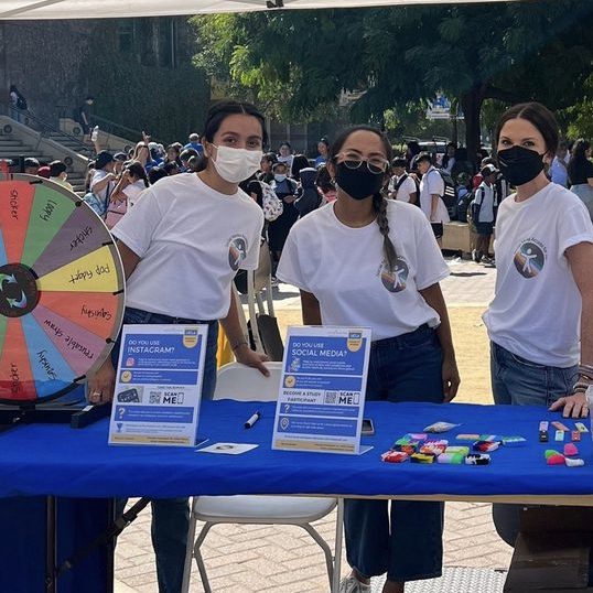 Three students with face masks on stand behind a bright blue desk to get participants to enter the study
