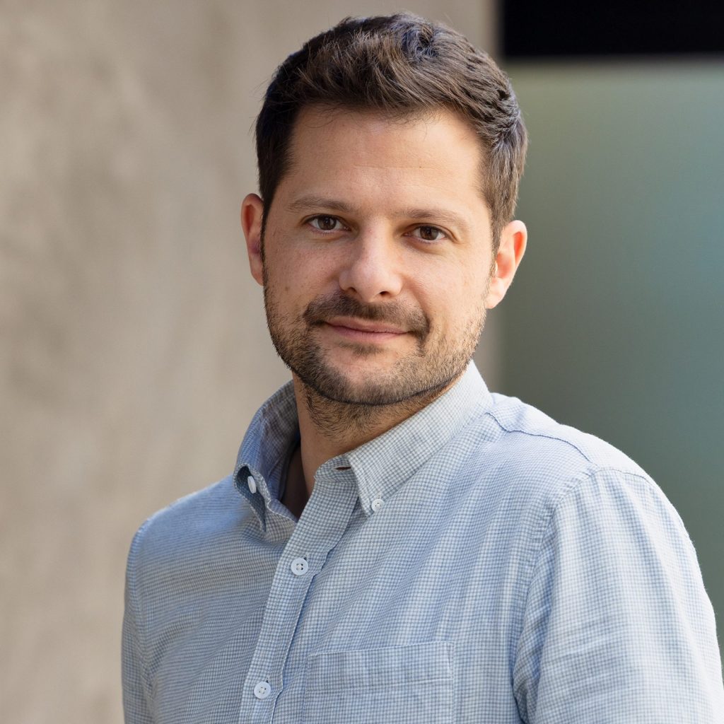 A white male in his 30s with short, dark hair, and stubble wearing a blue button up shirt against a gray/blue background.