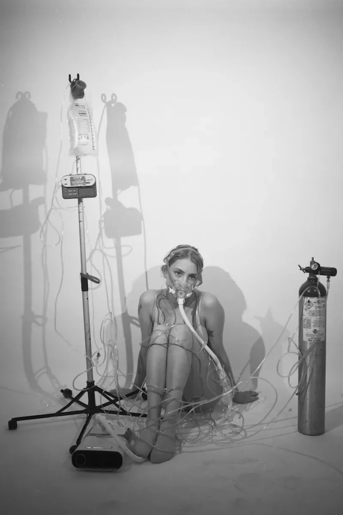 Black and white image of Rowan O'Bryan as part of her self-portrait project, "Invitation to Stare." Rowan is seen sitting wearing an oxygen mask with tubes wrapped around her body.