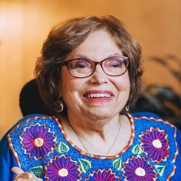 Headshot of Judith E. Heumann. She is a white woman with glasses smiling to the camera and wearing a bright blue shirt with purple flowers.