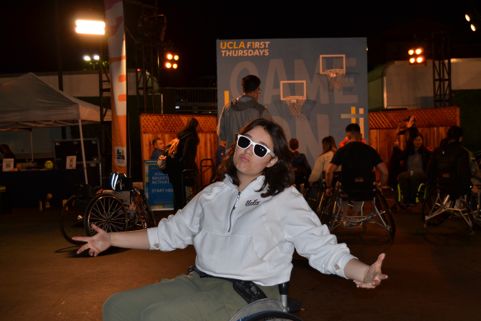 UCLA student Clarissa Lesky poses in front of the wheelchair basketball court. She is wearing a white UCLA sweater and sunglasses, and is sitting in a sports wheelchair with her arms spread out.