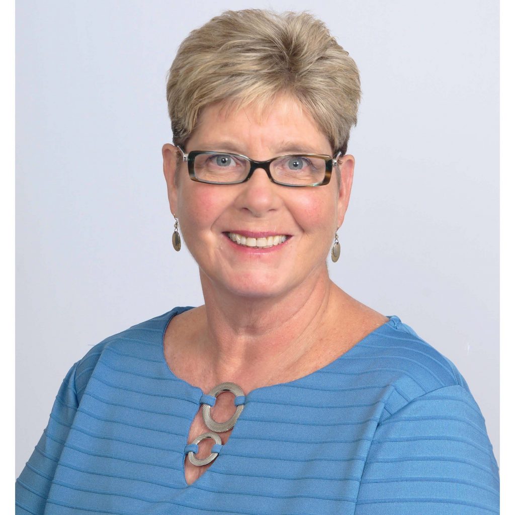 A headshot of Dr. Ammons smiling at the camera. She has short blonde hair and is wearing dark rimmed glasses and a light blue top.