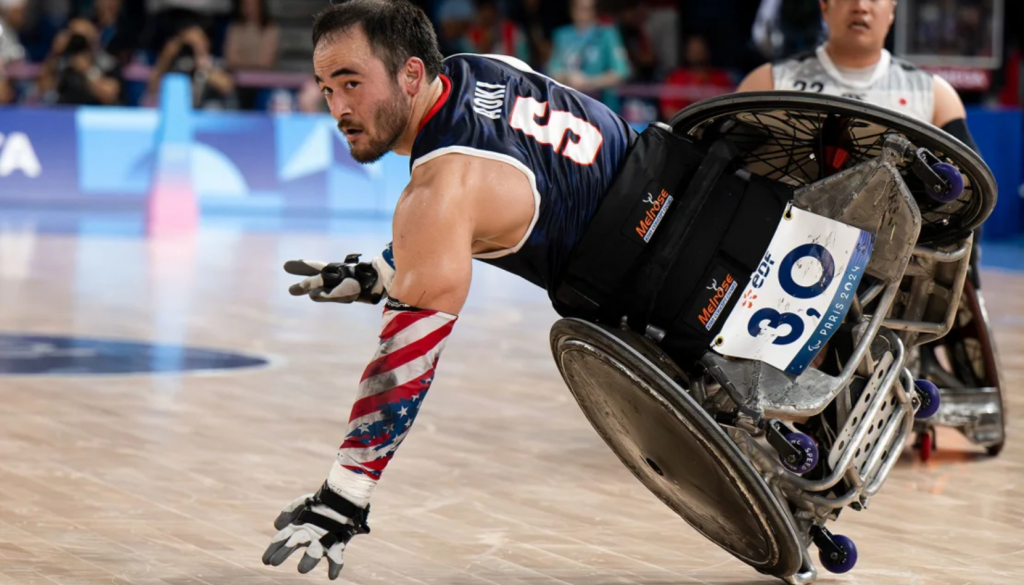 Chuck Aoki, a wheelchair rugby player wearing a dark blue jersey with the number 3, is in action on the court.