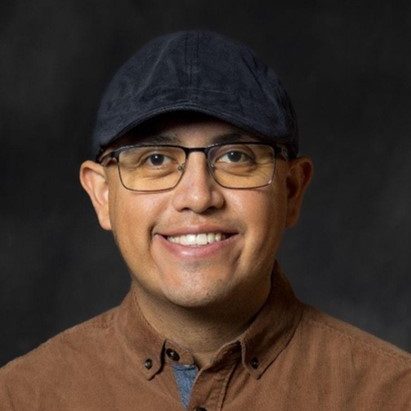 A young man wears glasses, a black cap, and brown shirt while smiling into the camera.