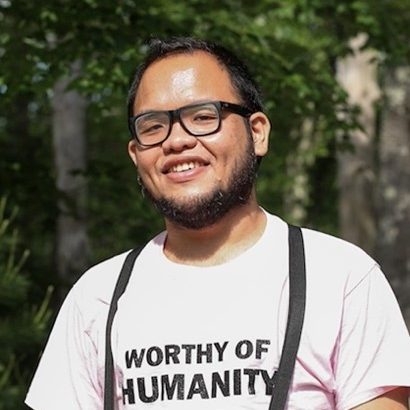 A male-presenting person with olive complexion, black hair and beard stands in front of trees. They are wearing glasses, suspenders, and a pink shirt with the text “Worthy of Humanity.”