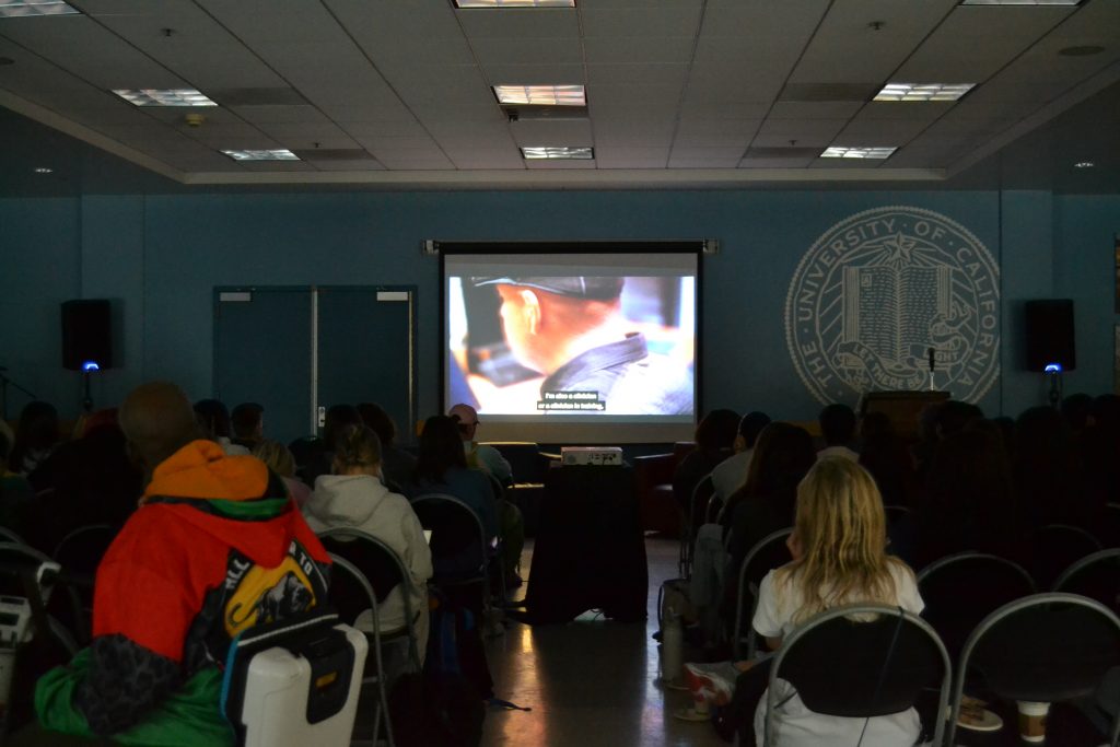 A dimly lit auditorium with an audience seated in rows, watching the film unseen on a screen at the front. The projection shows the back of a person wearing a cap with subtitles visible at the bottom of the screen. The University of California seal is faintly visible on the wall to the right.