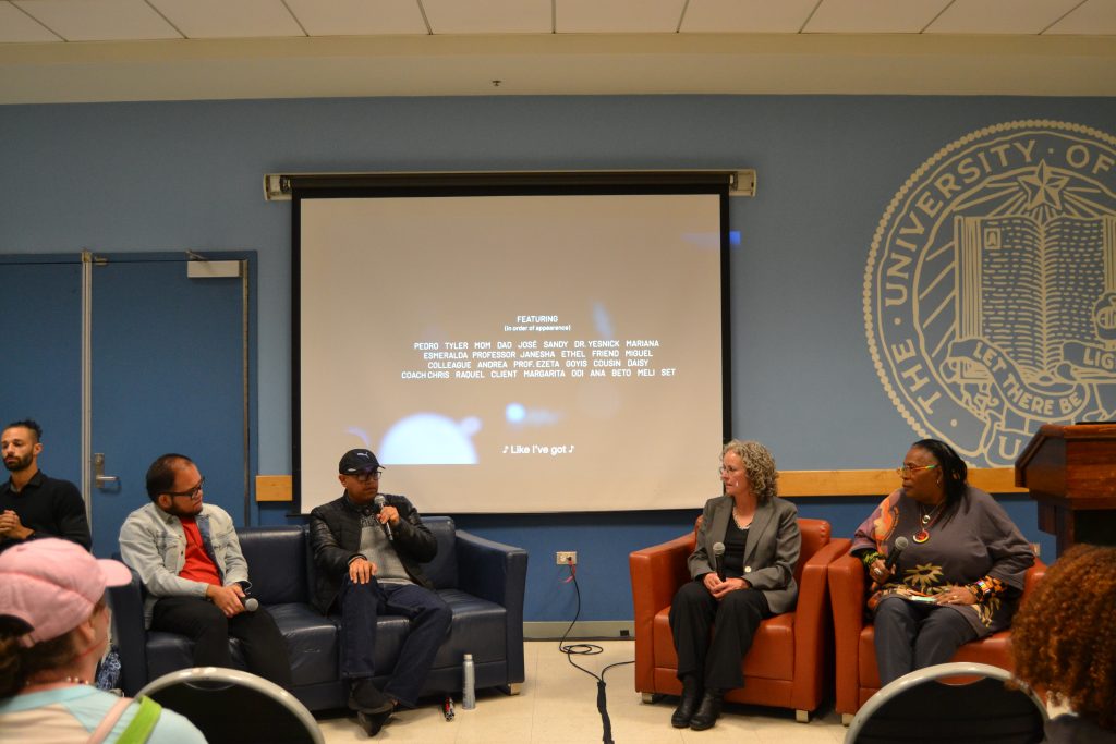 A panel discussion takes place in a room at the University of California. Four panelists and an interpreter sit on chairs in front of a projection screen displaying the end credits of unseen. The panelists hold microphones and engage with the audience. The University of California seal is visible on the wall to the right. Audience members are partially visible in the foreground.