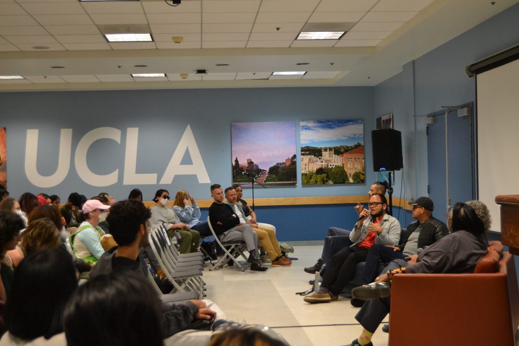 A panel discussion in a room with "UCLA" prominently displayed on a wall behind the audience. The audience is seated in rows of chairs, attentively listening to panelists seated on the right side of the image. The panelists are holding microphones and engaged in conversation. Large framed photos of the UCLA campus are visible on the wall in the background.