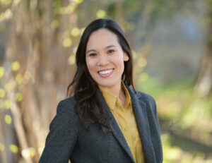 Dr. Duong has medium tan skin and long dark brown hair. She wears a yellow blouse with buttons and a checkered gray blazer. The background is a blurred mix of light and dark green hues