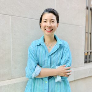 An Asian-American with black hair pulled back in a low bun smiles with her arms crossed. She is wearing a necklace strung with bright blue crystal gems and an azure blue button-down shirt. The background is of a building with light grey cemented walls.