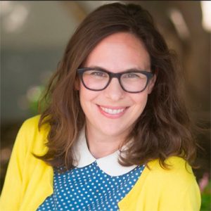 A photo of Sharrona Pearl with long brown hair smiling warmly at the camera. She is wearing black-framed glasses, a blue polka dot blouse with a white collar, and a bright yellow cardigan. The background is softly blurred with natural light.