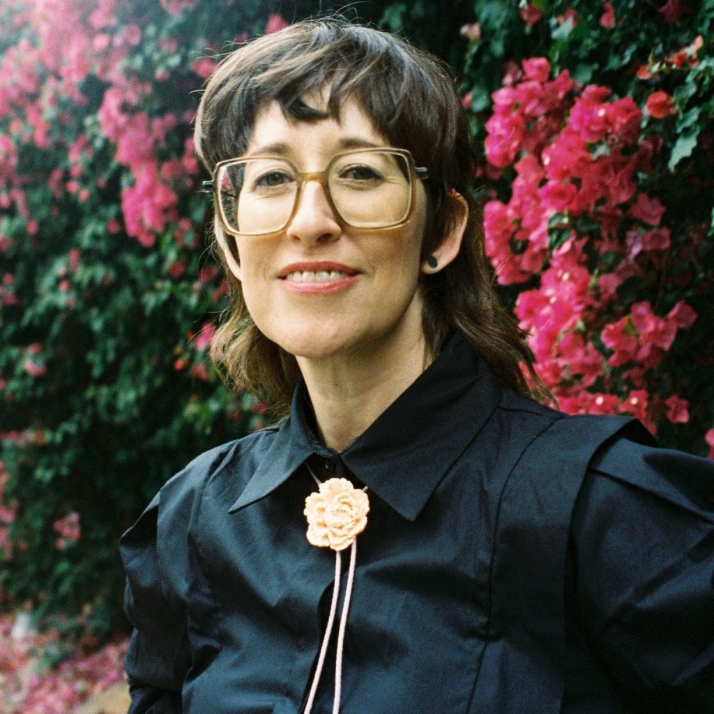 Alison, a smiling white woman with disheveled brown hair, wearing large square brown glasses, a black shirt and a peach flower bolo tie, stands in front of bright pink bougainvillea.