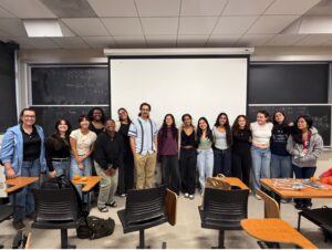 In a classroom setting, a group of fourteen female students are standing shoulder to shoulder smiling together. They are joined by their male professor, who is standing near the middle of the group. Most of the students are dressed casually in jeans paired with t-shirts or sweaters.