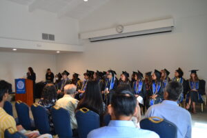 Dean Galván stands at the podium to the left of the graduating seniors. She is wearing a black pan suit.