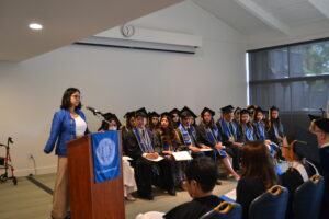 Quinn O'Connor stands at the podium to the left of the graduating seniors. Quinn is wearing a blue blazer with a white top underneath.