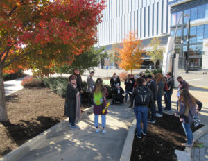 Photograph of one of the access audit groups (approximately 15 people) in Nashville. Everyone is wearing jackets and gathers around a small ramp surrounded by young trees with fall color. Alexa holds a yellow level to measure slope and Karen Braitmayer is visible in her wheelchair next to the ASL interpreter.