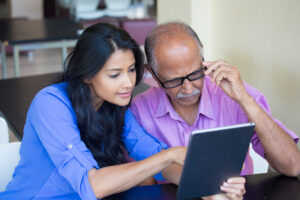 Woman in blue top helps man in purple top navigate on a tablet.