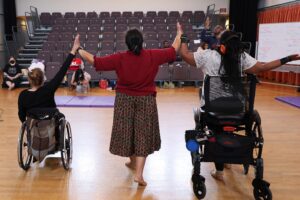 Three disabled dancers touching hands with arms raised; wheelchair user (left), person standing (center), and person standing with a walker.