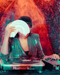 DJ Maria Chavez wears a green sequin blazer and stands behind a turntable as she holds up a broken record to cover her face. She stands in front of a red and black background.