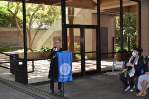 Chair Vic Marks stands at the podium delivering her opening remarks. She wears all black and stands in front of wall of windows. 