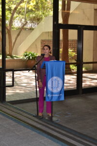 Dean Galván wears a monochromatic pink suit. She stands at the podium in front of a wall of windows. 