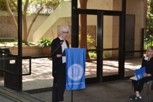 Former AVP Lucy Blackmar wears a black cardigan over a white top with black pants. She stands at the podium in front of a wall of windows. 