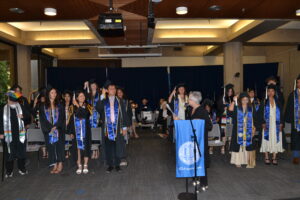 Chair Marks leads graduates in turning their tassels. Graduates stand at their seats behind her. 