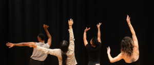 A group of dancers stand in front of a black wall. Each person is posed with their arms outstretched to the sky and side.
